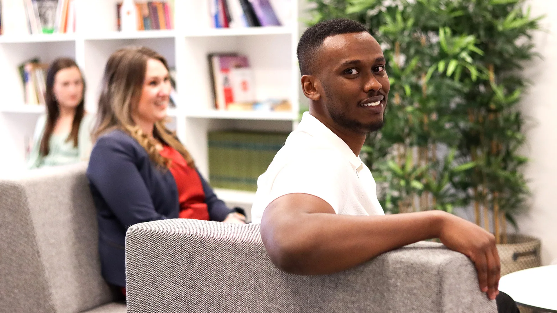 En man som sitter i en fåtölj ler in i kameran. I bakgrunden syns två kvinnor.