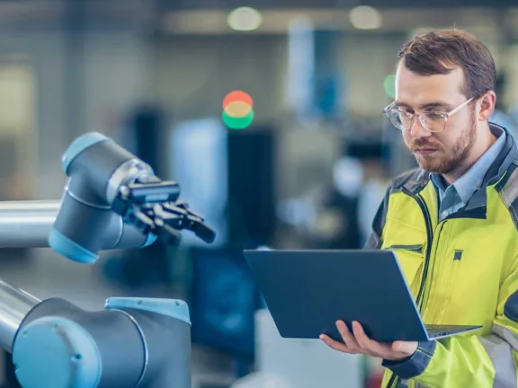 Man i arbetskläder som står vid en robot och tittar på sin laptop.
