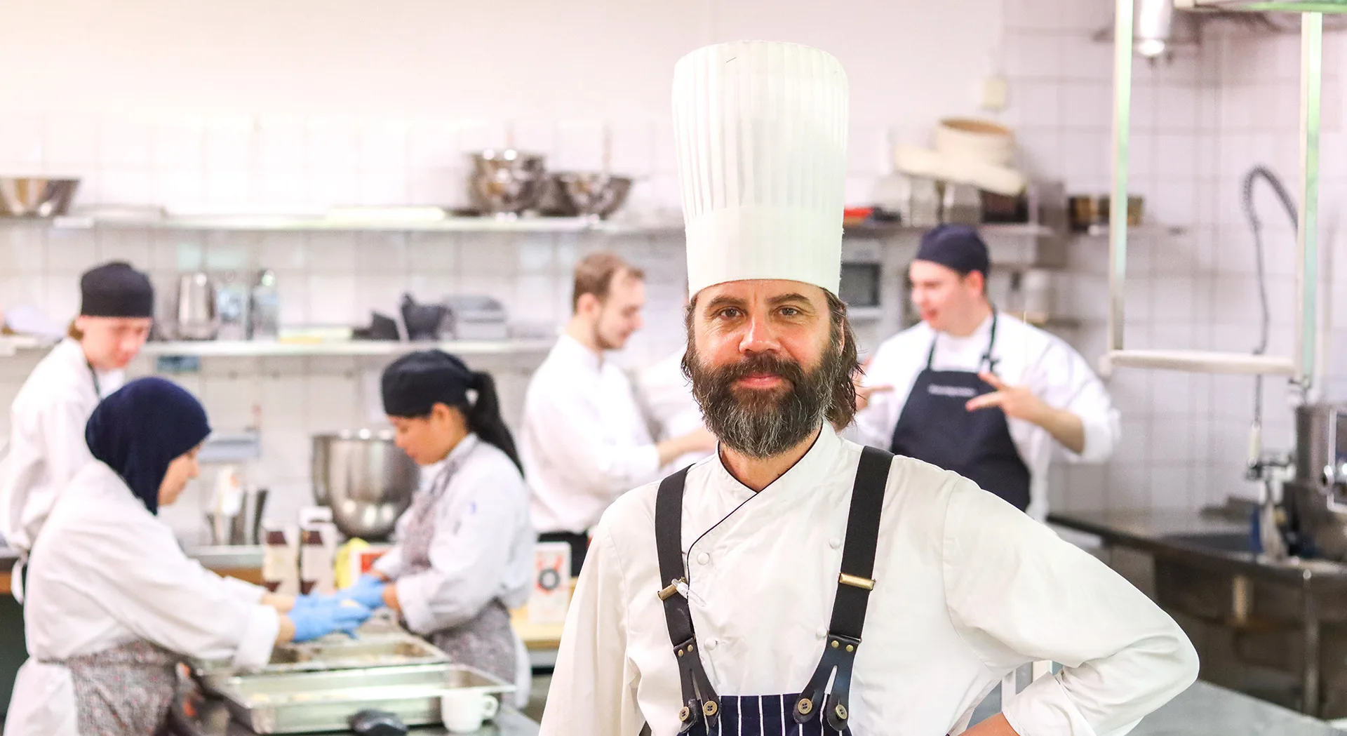 En manlig kock i ett storkök som tittar in i kameran. I bakgrunden syns kvinnor och män som arbetar.