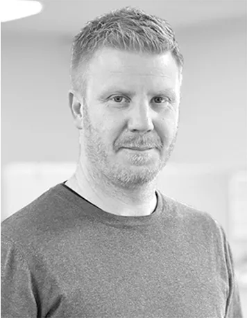 Svartvit porträttbild av Joakim Bramby som står i ett kontorslandskap på TUC Yrkeshögskola. Joakim har skäggstubb, ljust kortklippt hår. Han bär en grå finstickad tröja