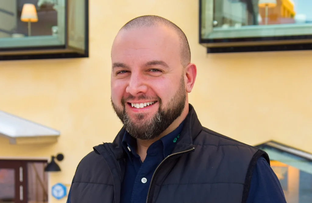 Anestis står utomhus och tittar in i kameran. Bakom honom finns en gul husfasad med två fönster och en dörr.