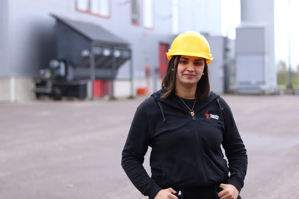 Zainab står framför ett kraftvärmeverk och tittar in i kameran. Hon har på sig svarta arbetarkläder och en gul skyddshjälp. Hon har händerna i sidan och ser bestämd ut.