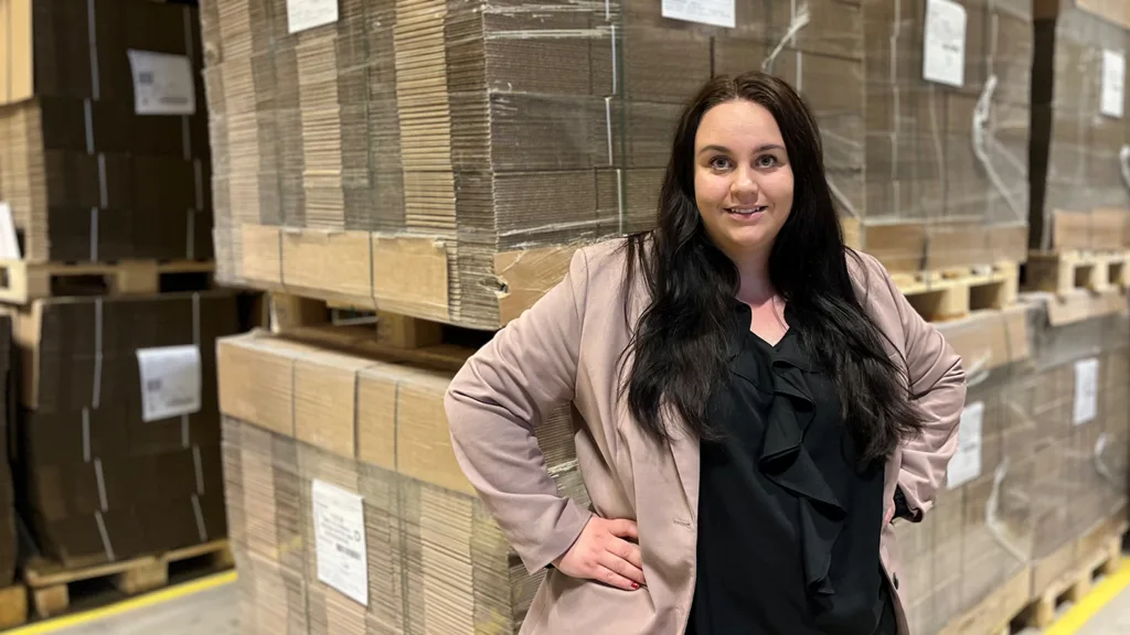 Mikaela Melin står i vänstra delen av bilden och har händerna på sina höfter. Hon tittar in i kameran och ler. I bakgrunden syns en lagerlokal med lastpallar fyllda med flyttkartonger.