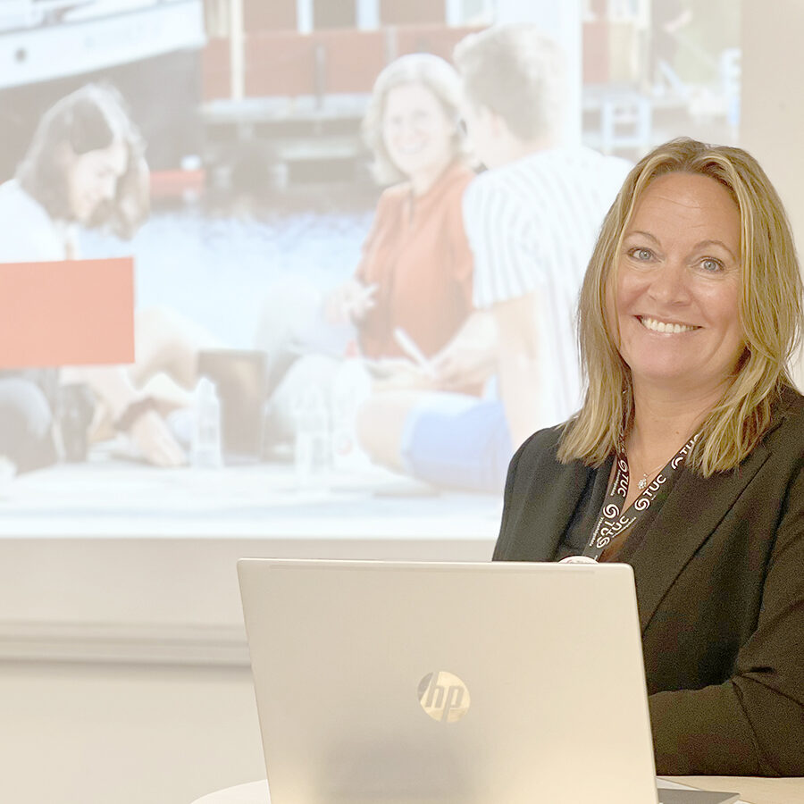 Utbildningskonsulten Malin Swenson står i ett klassrum vid ett stå bord. På bordet står en öppen laptop och bakgrunden ser man en projicerad powerpoint-presentation.