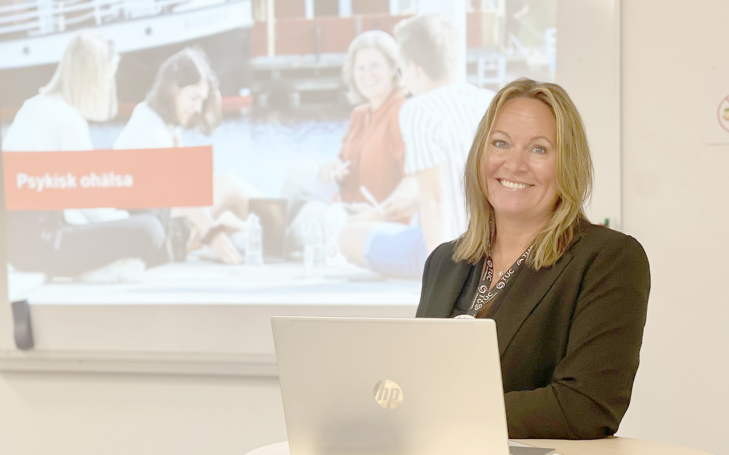 Utbildningskonsulten Malin Swenson står i ett klassrum vid ett stå bord. På bordet står en öppen laptop och bakgrunden ser man en projicerad powerpoint-presentation.