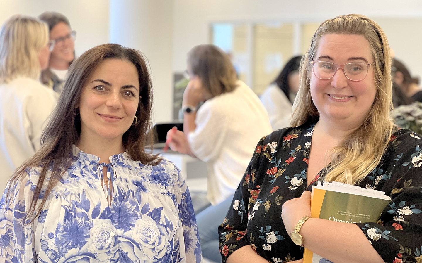 Yazi Al-Yousifi och Jennifer Rae står i förgrunden och ler mot kameran. Jennifer håller i en kursbok. I bakgrunden ses de andra studerande som har en paus i studierna