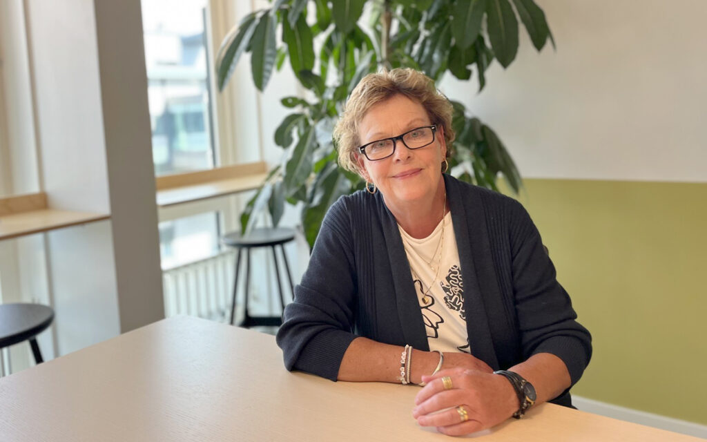 Åsa Stenman sitter vid ett bord i ljust träslag. I bakgrunden syns ett fönsterparti och en stor grön växt. Åsa är klädd i en mörkblå kofta och ljus t-shirt. Hon har kort hår och bär fyrkantiga glasögon med mörk båge.