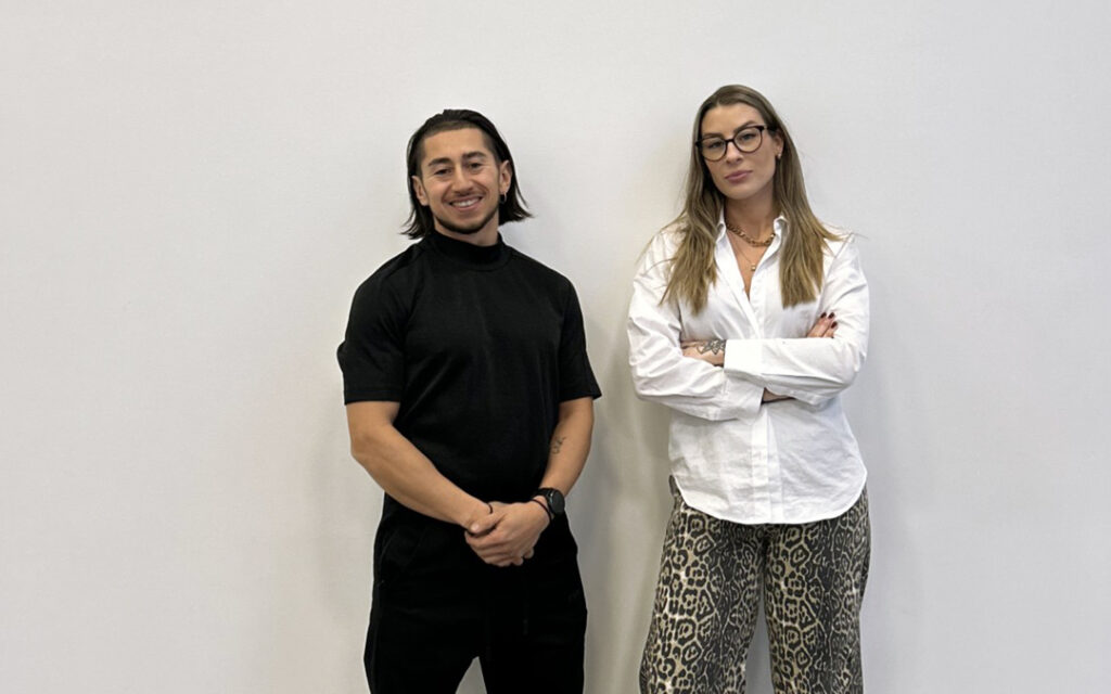 Johannes Ojanperä och Amanda Sörensen poserar framför en vit vägg. Johannes har mörkt axellångt hår och klädd helt i svart. Amanda har långt, brunt hår, bär mörka glasögon och en vit skjorta med mörka byxor.