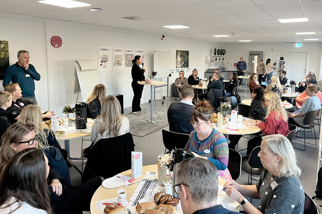 På bilden syns ett stort sällskap personer som sitter runt flera runda bord. På borden finns frukostfrallor och kaffekoppar. I vänstra hörnet av bilden står Cecilia Mårtensson som är verksamhetsansvarig på TUC i Linköping och pratar med personerna som sitter runt borden