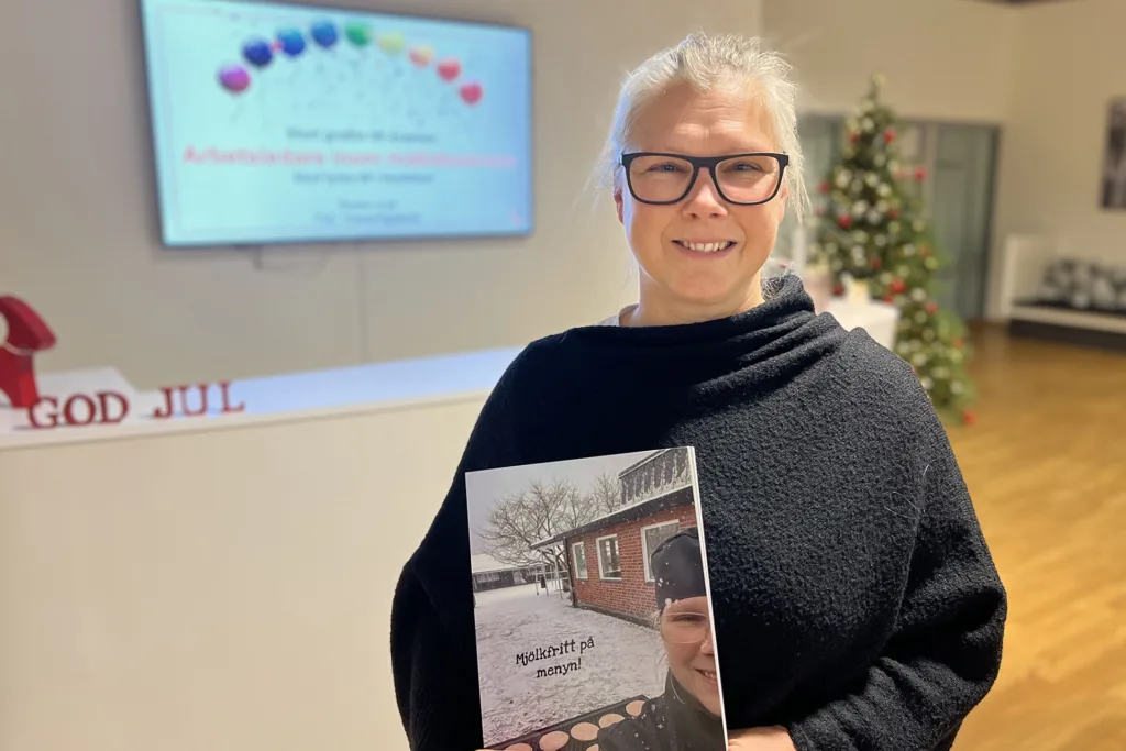 Jennie står i TUCs lokaler där hon håller en kokbok med mjölkallergianpassade recept. Hon bär en svart ponch, har grått långt hår uppsatt i en tofs. Hon ser glad ut.