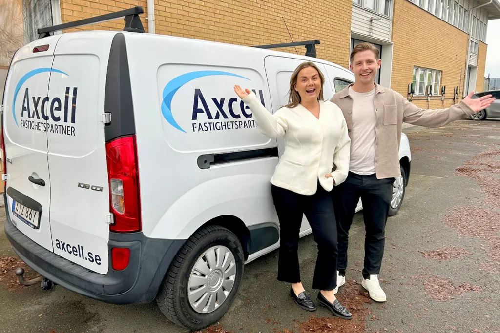Linnea och Gustav står framför en vit skåpbil med texten Axcell Fastighetspartner. De ser glada ut och håller ut handen som i en vinkning