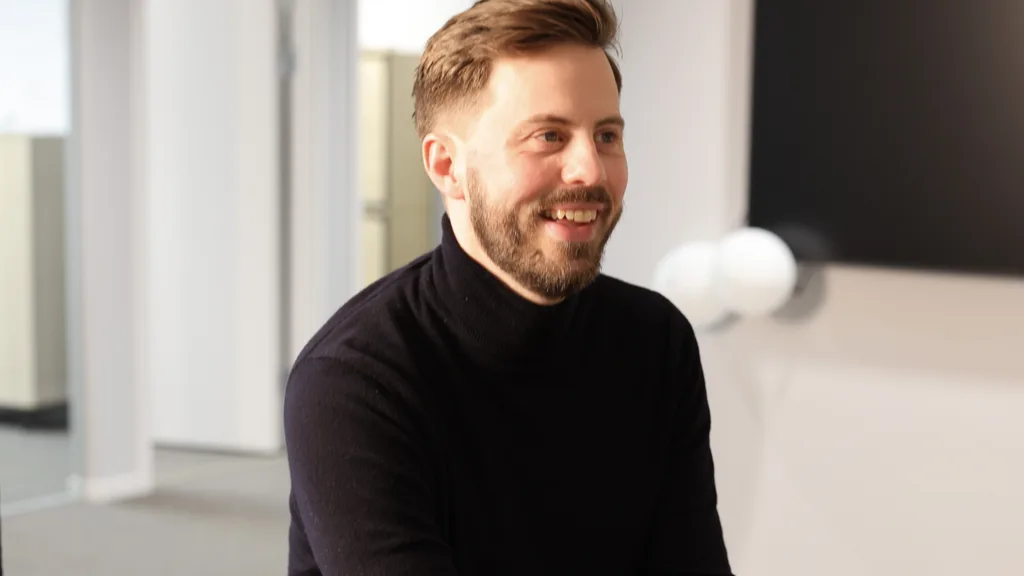 Jacob Linusson sitter i ett öppet kontorslandskap. Han har kort, brunt hår och kortklippt skägg. Han bär en svart polotröja. Jacob ler och tittar åt höger.