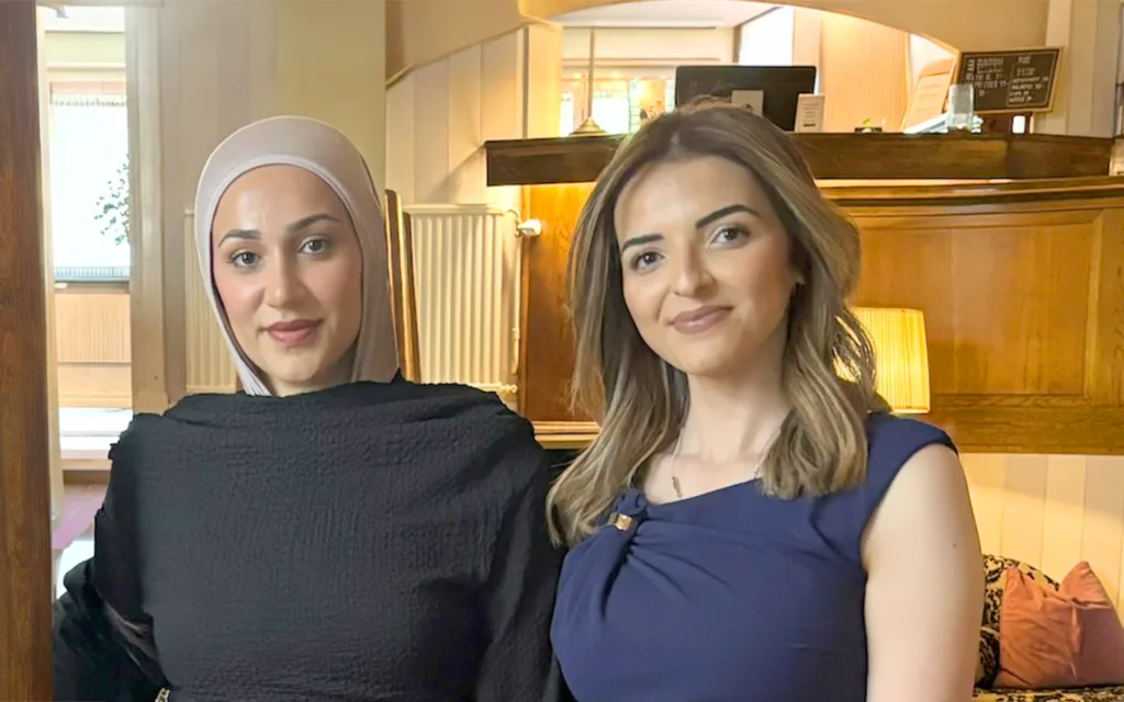 Narjes Hashem och Roula Enegård står tillsammans i en hotellobby. Narjes bär en svart klänning och beige slöja medan Roula har en blå klänning.