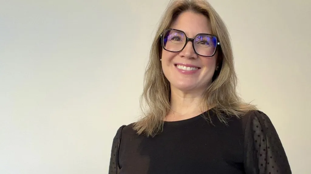 Porträttbild Nicolina som står mot en ljus bakgrund. Hon har axellångt hår, svarta markerade glasögon och en svart topp. Hon ser in i kameran och ser glad ut.