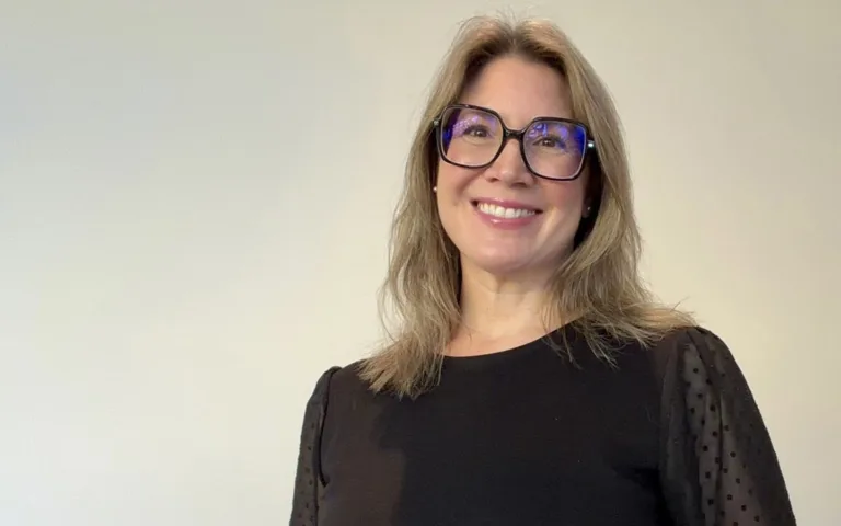 Porträttbild Nicolina som står mot en ljus bakgrund. Hon har axellångt hår, svarta markerade glasögon och en svart topp. Hon ser in i kameran och ser glad ut.