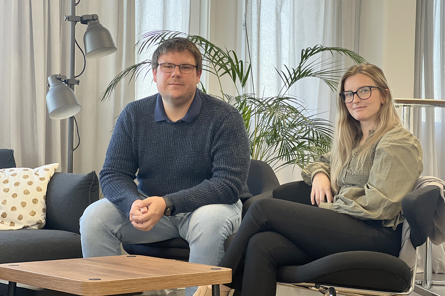 Kristoffer och Felicia sitter i en mörk soffgrupp och tittar in i kameran. Kristoffer har mörkblå tröja och ljusa jeans medan Felicia har en olivgrön tunika och svarta byxor.