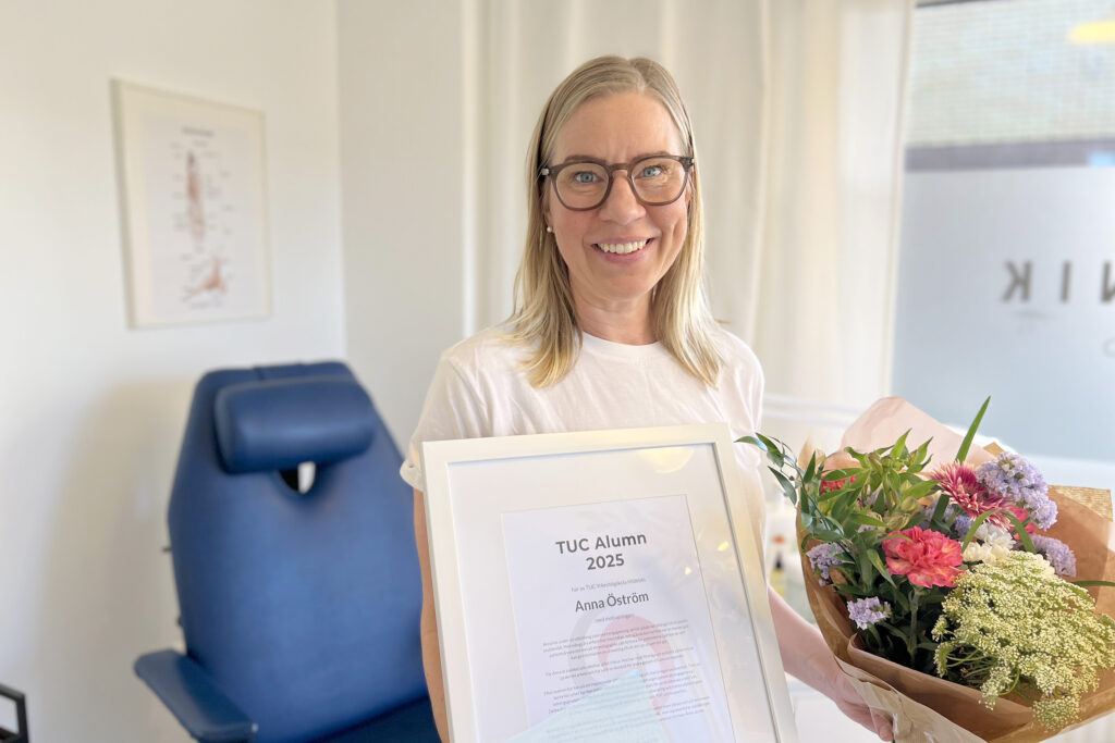 I bilden står Anna i sin fotklinik. i Bakgrunden syns en blå behandlingsstol. Anna håller sitt diplom för Årets Alumn i högra handen och i vänstra har hon en blomsterbukett. Hon ser glad ut.
